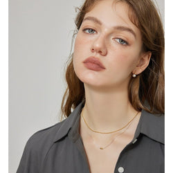 Tweevoudige zilveren sieraden ketting met gouden boon-accent, gedragen door vrouw met bruin haar in witte top. Minimalistische gelaagde kettingen met rijstkralen patroon, elegant liggend rond de hals voor moderne mode-accessoire.