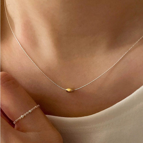 Stijlvolle zilveren ketting met gouden boon-accent, gedragen door vrouw in witte top, een trendy en minimalistisch mode-accessoire.