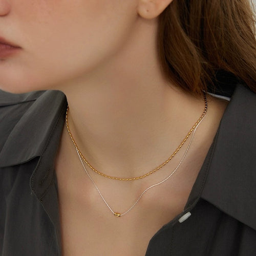 Set van twee slanke zilveren kettingen; één met een gouden boon en één met rijstvormige kralen; geschikt voor afzonderlijk dragen of stijlvolle gelaagdheid.