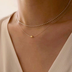 Tweevoudige zilveren ketting met gouden boon-accent, gedragen door vrouw met bruin haar in een witte top, voor een elegante, moderne look