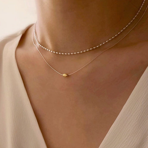 Tweevoudige zilveren ketting met gouden boon-accent, gedragen door vrouw met bruin haar in een witte top, voor een elegante, moderne look
