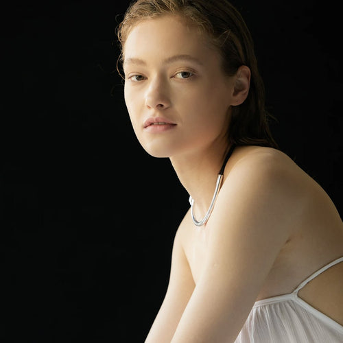 Een vrouw in een witte jurk zit op een tafel, met een elegante zilveren ketting en leren touwtje om haar hals.