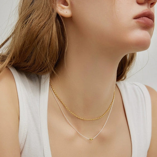 Tweevoudige zilveren sieraden ketting met gouden boon-accent, gedragen door vrouw met bruin haar in witte top. Minimalistische gelaagde kettingen met rijstkralen patroon, elegant liggend rond de hals voor moderne mode-accessoire.